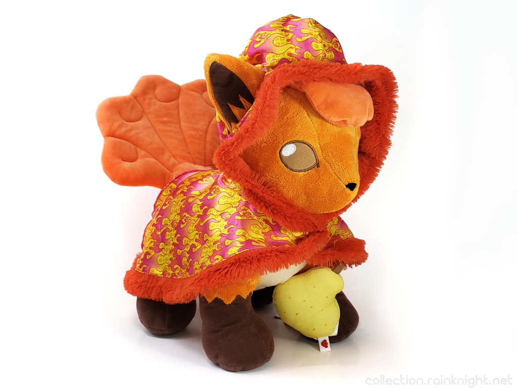 Build-A-Bear Vulpix Plush wearing a shiny cape with red and yellow fire imagery and a furry led-orange lining. It's holding a Sitrus berry, which looks kind of like a pear.